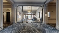 This distinguished single-floor space showcases a seamless blend of classical architectural heritage and contemporary design. The expansive lobby and hallway areas are unified by richly patterned, dark marble-effect flooring that flows continuously throughout, complemented by original ornate plasterwork detailing on the high ceilings, decorative corbels, and columned archways that speak to the building's historic character.

A striking steel-framed, floor-to-ceiling glass partition wall defines the main office area, offering full visual transparency while maintaining spatial separation. Inside, natural light pours through large arched transom windows and traditional timber-framed doors, illuminating a well-appointed workspace furnished with conference and workstation setups. A bold geometric pendant light fixture adds a modern focal point overhead.

The interconnecting hallways are generous in scale, lined with ornamental columns and detailed crown molding. Comfortable seating areas with plush upholstered chairs and marble-topped tables are thoughtfully positioned throughout. A staircase with marble-clad risers leads further into the building, flanked by an elevator and a traditional mailbox cabinet. The overall ambiance is one of refined elegance, balancing historic grandeur with polished, contemporary functionality.