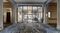 This distinguished single-floor space showcases a seamless blend of classical architectural heritage and contemporary design. The expansive lobby and hallway areas are unified by richly patterned, dark marble-effect flooring that flows continuously throughout, complemented by original ornate plasterwork detailing on the high ceilings, decorative corbels, and columned archways that speak to the building's historic character.

A striking steel-framed, floor-to-ceiling glass partition wall defines the main office area, offering full visual transparency while maintaining spatial separation. Inside, natural light pours through large arched transom windows and traditional timber-framed doors, illuminating a well-appointed workspace furnished with conference and workstation setups. A bold geometric pendant light fixture adds a modern focal point overhead.

The interconnecting hallways are generous in scale, lined with ornamental columns and detailed crown molding. Comfortable seating areas with plush upholstered chairs and marble-topped tables are thoughtfully positioned throughout. A staircase with marble-clad risers leads further into the building, flanked by an elevator and a traditional mailbox cabinet. The overall ambiance is one of refined elegance, balancing historic grandeur with polished, contemporary functionality.