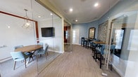 This single-floor space is thoughtfully designed to accommodate a variety of social and professional needs. A standout feature is the glass-walled conference room, which provides a sense of openness while offering a defined, private area for meetings. Inside, a round wooden table paired with contemporary teal chairs creates an inviting setting, complemented by a wall-mounted flat-screen television. The sliding glass partitions allow the space to be reconfigured as needed, adding functional flexibility.

Adjacent to the enclosed room, an open lounge area is finished in a calming blue-gray tone, providing a welcoming contrast to the neutral carpeted flooring that runs throughout. This area features high-top bar tables with matching dark-framed stools upholstered in teal, creating a casual yet polished atmosphere suitable for informal gatherings or individual use.

Crown molding along the ceiling adds a refined architectural detail, while recessed lighting ensures consistent, well-distributed illumination throughout. Decorative accents, including a geometric mirror and a colorful abstract artwork, contribute personality and visual interest to the space. A frosted-glass door provides access to an adjoining area, maintaining natural light flow while preserving privacy.