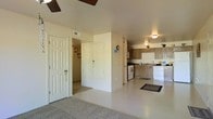 This single-floor residence offers a practical and open layout that connects the living areas with ease. The main living space features light-colored flooring and carpeted sections, creating a comfortable and welcoming environment. A ceiling fan is mounted overhead, contributing to the room's ventilation and casual ambiance.

The kitchen is positioned at the rear of the open-plan area and is equipped with a gas range, a white refrigerator, and a dishwasher. Medium-toned wood cabinetry lines the walls, providing ample storage, while a sink sits beneath additional upper cabinets. The kitchen flows naturally into the surrounding living space, making it ideal for everyday use.

Adjacent to the main area, a hallway leads to additional rooms, with a pair of white panel doors visible — one appearing to serve as a closet and another leading to a secondary space. The transition between the carpeted and hard-floor zones helps define separate functional areas within the open floor plan.

Natural and artificial lighting work together to keep the interior bright throughout the day. The overall design emphasizes functionality and straightforward living, with a clean, neutral color palette that keeps the space feeling airy and easy to personalize.