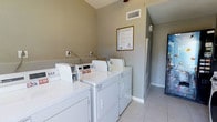 This shared laundry room is a practical, well-organized facility designed to meet residents' laundry needs. The room features a row of white top-loading washing machines paired with matching dryers, all equipped with digital controls for ease of use. The machines are neatly arranged along one wall, providing an efficient layout that maximizes the available space.

The room is finished with neutral taupe-toned walls and clean light-colored tile flooring, creating a bright and tidy environment. A ceiling fan is mounted overhead, promoting air circulation throughout the space. An HVAC vent ensures additional climate control, keeping the room comfortable year-round.

A framed informational notice is posted on the wall, providing residents with usage guidelines and facility details. Electrical outlets are accessible along the wall, offering added convenience. A vending machine is partially visible near the entrance, and a utility door provides access to adjacent areas.

The overall design is functional and straightforward, prioritizing accessibility and efficiency. The well-maintained condition of the equipment and the clean, orderly layout make this laundry room a reliable and convenient shared amenity.