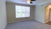 This single-floor residence offers a comfortable and functional layout designed with classic finishes throughout. The interior is finished with soft wall-to-wall carpeting that adds warmth underfoot, complemented by neutral-toned walls that create a calm, versatile backdrop for any furnishing style. Crown molding runs along the ceiling line, lending a polished, traditional character to the space. A ceiling fan with a built-in light fixture provides both overhead illumination and year-round climate comfort. The room is anchored by a large picture window dressed with horizontal blinds, allowing for adjustable natural light and privacy. The window frames a verdant outdoor view of mature trees and lush greenery, bringing a sense of the outdoors inside and creating a bright, airy atmosphere. White baseboards provide a crisp contrast against the flooring and walls, reinforcing the clean, well-maintained aesthetic. The overall ambiance is quiet and inviting, with thoughtful details that reflect a move-in-ready condition. The single-floor layout ensures easy accessibility and seamless flow throughout the home, making it a practical and pleasant living environment.