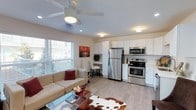This single-floor space features an open-concept layout that seamlessly connects the living area and kitchen, creating a functional and cohesive environment. The living area is anchored by a comfortable sectional sofa, a glass-top coffee table, and a cowhide-style area rug resting on light-toned laminate flooring that extends throughout the space. A large window with horizontal blinds allows generous natural light to fill the room, while a ceiling fan with an integrated light fixture provides both airflow and overhead illumination.

The kitchen is outfitted with white shaker-style cabinetry, light countertops, and a full suite of stainless steel appliances, including a refrigerator, electric range, and over-the-range microwave. The clean, neutral palette of the kitchen complements the overall brightness of the space.

A built-in desk area with upper cabinetry is tucked neatly between the living and kitchen zones, offering a compact and dedicated workspace without occupying additional floor area. Recessed lighting supplements the natural light throughout. The overall ambiance is modern, clean, and efficiently designed.