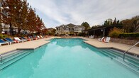 Located at 8718 Wintersweet Ln in Charlotte, NC, this outdoor amenity space offers a thoughtfully designed retreat for residents and guests alike. At the heart of the area is a generously sized, resort-style swimming pool with crystal-clear blue water, surrounded by a spacious concrete pool deck furnished with lounge chairs and Adirondack-style seating, providing ample room for relaxation and recreation.

Adjacent to the pool is a covered pergola patio, constructed with a sleek dark metal overhead lattice structure supported by substantial columns. The patio floor features classic cobblestone pavers, lending a refined, textured aesthetic to the space. Beneath the pergola, comfortable seating arrangements — including lounge chairs and dining sets with blue accent chairs — invite casual outdoor gatherings and al fresco dining.

A low-profile stone retaining wall frames the patio perimeter, complemented by lush, mature greenery including tall evergreen trees and climbing vines that add natural privacy and visual warmth. Wrought iron fencing elegantly defines the boundary of the space while maintaining open sightlines to the surrounding landscape.

The multi-story residential buildings in the background, clad in blue and gray siding with stone accents, provide an attractive architectural backdrop. Overall, this outdoor amenity area combines functional design with inviting aesthetics, creating an ideal communal space for year-round enjoyment in Charlotte's Ballantyne area.
