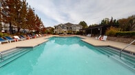 Located at 8718 Wintersweet Ln in Charlotte, NC, this outdoor amenity space offers a thoughtfully designed retreat for residents and guests alike. At the heart of the area is a generously sized, resort-style swimming pool with crystal-clear blue water, surrounded by a spacious concrete pool deck furnished with lounge chairs and Adirondack-style seating, providing ample room for relaxation and recreation.

Adjacent to the pool is a covered pergola patio, constructed with a sleek dark metal overhead lattice structure supported by substantial columns. The patio floor features classic cobblestone pavers, lending a refined, textured aesthetic to the space. Beneath the pergola, comfortable seating arrangements — including lounge chairs and dining sets with blue accent chairs — invite casual outdoor gatherings and al fresco dining.

A low-profile stone retaining wall frames the patio perimeter, complemented by lush, mature greenery including tall evergreen trees and climbing vines that add natural privacy and visual warmth. Wrought iron fencing elegantly defines the boundary of the space while maintaining open sightlines to the surrounding landscape.

The multi-story residential buildings in the background, clad in blue and gray siding with stone accents, provide an attractive architectural backdrop. Overall, this outdoor amenity area combines functional design with inviting aesthetics, creating an ideal communal space for year-round enjoyment in Charlotte's Ballantyne area.