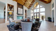This single-story amenity clubhouse is a masterclass in modern design, combining warm natural materials with contemporary architectural elements. The centerpiece of the space is a grand great room featuring soaring vaulted ceilings clad in rich wood tongue-and-groove paneling, supported by exposed beams and illuminated by sculptural ring chandeliers. A dramatic floor-to-ceiling white stone fireplace anchors one wall, flanked by built-in shelving and a wall-mounted display, while expansive floor-to-ceiling glass windows on the opposing wall flood the interior with natural light and frame panoramic views of the outdoor pool area and surrounding community.

The lounge area offers multiple seating arrangements with a curated mix of upholstered sofas, accent chairs, and stylish area rugs over wide-plank hardwood and large-format tile flooring. A dedicated kitchen bar area features a gas cooktop island with a warm quartz countertop, gray cabinetry, open glass shelving, and a textured tile backsplash. A corridor extends the space further with additional casual seating nooks, game tables, and banquette-style seating, all bathed in abundant natural light from clerestory and full-height windows throughout.