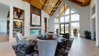 This single-story amenity clubhouse is a masterclass in modern design, combining warm natural materials with contemporary architectural elements. The centerpiece of the space is a grand great room featuring soaring vaulted ceilings clad in rich wood tongue-and-groove paneling, supported by exposed beams and illuminated by sculptural ring chandeliers. A dramatic floor-to-ceiling white stone fireplace anchors one wall, flanked by built-in shelving and a wall-mounted display, while expansive floor-to-ceiling glass windows on the opposing wall flood the interior with natural light and frame panoramic views of the outdoor pool area and surrounding community.

The lounge area offers multiple seating arrangements with a curated mix of upholstered sofas, accent chairs, and stylish area rugs over wide-plank hardwood and large-format tile flooring. A dedicated kitchen bar area features a gas cooktop island with a warm quartz countertop, gray cabinetry, open glass shelving, and a textured tile backsplash. A corridor extends the space further with additional casual seating nooks, game tables, and banquette-style seating, all bathed in abundant natural light from clerestory and full-height windows throughout.