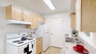 This single-floor residence offers a well-organized layout across two bedrooms, a living room, a hallway, and a utility room. The open living area features neutral wall-to-wall carpeting and clean white walls, creating a bright and welcoming atmosphere. A sliding glass door leads to an outdoor balcony, filling the room with natural light and providing easy access to the exterior. The front entry door is conveniently located within the living space. The galley-style kitchen is efficiently designed with light wood cabinetry, granite-look countertops, and a full suite of white appliances including an electric range and full-size refrigerator. A skylight-style overhead fixture brightens the kitchen, which also features tile flooring and a double stainless steel sink. The generously sized primary bedroom offers plush carpeting and is one of the more spacious rooms in the home. The secondary bedroom also features carpet flooring and a window with outdoor views, making it a comfortable and functional space. Both rooms are finished with crisp white walls and clean trim throughout. A hallway connects the main living areas to the bedrooms, while a dedicated utility room adds practical storage and functional convenience to this well-appointed residence.
