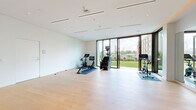 This thoughtfully designed fitness studio offers a bright and open workout environment defined by clean, contemporary architecture. Light hardwood flooring spans the entire space, complementing the crisp white walls and a smooth ceiling fitted with recessed lighting and integrated linear air vents for a polished, minimalist aesthetic.

A full-length wall mirror on one side of the room enhances the sense of openness, making the space feel expansive and ideal for a range of exercise activities. The studio is well-equipped with cardio machines, including stationary bikes and a stair climber, as well as a dedicated storage area housing dumbbells, weight plates, resistance equipment, and stability balls — all neatly organized along a built-in bench nook.

The standout architectural feature is the floor-to-ceiling folding glass door system that opens the studio entirely to a private outdoor terrace. The terrace is finished with lush green turf, manicured landscaping, and ornamental plantings, creating a seamless indoor-outdoor connection. From this vantage point, sweeping views of the city skyline are visible, adding an inspiring backdrop to any workout. Frosted glass interior doors provide access to adjacent spaces, completing this premium, self-contained fitness environment.