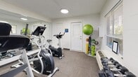 Located at 401 W Park Ave in Tallahassee, Florida, this fitness center is a efficiently organized single-floor facility designed to support a well-rounded workout experience. The main exercise area is equipped with a treadmill, elliptical trainer, and stationary bike, providing a solid range of cardio options. A rack of dumbbells lines one wall, complementing the cardio equipment with free-weight training capabilities. Additional exercise accessories, including a stability ball, medicine balls, resistance bands, and a jump rope, are neatly organized on a dedicated storage rack, maximizing the use of the available space. A large wall-mounted mirror spans one side of the room, visually expanding the space and allowing users to monitor their form during workouts. The flooring is covered in durable, low-pile carpet suited for fitness use. Recessed flush-mount ceiling lights provide consistent, even illumination throughout the space. A window with horizontal blinds allows natural light to filter in while offering views of the surrounding greenery. Climate control is handled by a wall-mounted PTAC unit. The facility also includes a private office area and a closet for additional storage. A bold blue entry door provides secure, designated access directly from the outdoors.