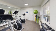 Located at 401 W Park Ave in Tallahassee, Florida, this fitness center is a efficiently organized single-floor facility designed to support a well-rounded workout experience. The main exercise area is equipped with a treadmill, elliptical trainer, and stationary bike, providing a solid range of cardio options. A rack of dumbbells lines one wall, complementing the cardio equipment with free-weight training capabilities. Additional exercise accessories, including a stability ball, medicine balls, resistance bands, and a jump rope, are neatly organized on a dedicated storage rack, maximizing the use of the available space. A large wall-mounted mirror spans one side of the room, visually expanding the space and allowing users to monitor their form during workouts. The flooring is covered in durable, low-pile carpet suited for fitness use. Recessed flush-mount ceiling lights provide consistent, even illumination throughout the space. A window with horizontal blinds allows natural light to filter in while offering views of the surrounding greenery. Climate control is handled by a wall-mounted PTAC unit. The facility also includes a private office area and a closet for additional storage. A bold blue entry door provides secure, designated access directly from the outdoors.
