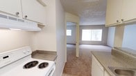 This single-floor space showcases a practical and open layout connecting the kitchen directly to the living area. The kitchen is equipped with a white electric coil-burner range and overhead ventilation hood, complemented by speckled laminate countertops on both sides that provide ample preparation space. Light-toned cabinetry lines the kitchen walls, contributing to a clean, bright atmosphere. The flooring in the kitchen area transitions from smooth tile to wall-to-wall carpeting as it opens into the adjacent living space. A wide sliding glass window or door at the far end of the living area invites natural light deep into the unit, creating a noticeably airy feel throughout. An additional window is visible in the adjoining room or hallway, further enhancing brightness. The textured ceilings are consistent throughout the visible areas, adding a classic residential character. The open passage between the kitchen and living room ensures easy flow and connectivity between the spaces. Overall, the layout is straightforward and functional, suited for everyday living with a neutral color palette of beige and white tones throughout.