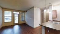 This single-floor residence offers a thoughtfully designed layout that balances comfort and functionality. The main living area immediately draws the eye with its rich, dark hardwood flooring that extends throughout the open-concept space, creating a warm and inviting ambiance. Natural light streams in through a glass-paneled front door and an adjacent window fitted with blinds, illuminating the room and offering views of the outdoors. The living area flows seamlessly into the kitchen, which features granite-style countertops and a stylish pendant light that adds a modern touch overhead. A partial wall divides the spaces while maintaining an open, airy feel. The neutral, warm-toned walls throughout complement the overall design and provide a versatile backdrop for any décor style. A dedicated laundry area adds everyday convenience, complete with a top-loading washing machine and a wall-mounted wire shelving unit above, maximizing vertical storage. A separate dryer hookup is also present. The space benefits from practical details such as built-in HVAC vents and ample outlet placements. Overall, this residence combines clean design with functional living spaces, making it a comfortable and efficient home.