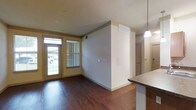 This single-floor residence offers a thoughtfully designed layout that balances comfort and functionality. The main living area immediately draws the eye with its rich, dark hardwood flooring that extends throughout the open-concept space, creating a warm and inviting ambiance. Natural light streams in through a glass-paneled front door and an adjacent window fitted with blinds, illuminating the room and offering views of the outdoors. The living area flows seamlessly into the kitchen, which features granite-style countertops and a stylish pendant light that adds a modern touch overhead. A partial wall divides the spaces while maintaining an open, airy feel. The neutral, warm-toned walls throughout complement the overall design and provide a versatile backdrop for any décor style. A dedicated laundry area adds everyday convenience, complete with a top-loading washing machine and a wall-mounted wire shelving unit above, maximizing vertical storage. A separate dryer hookup is also present. The space benefits from practical details such as built-in HVAC vents and ample outlet placements. Overall, this residence combines clean design with functional living spaces, making it a comfortable and efficient home.