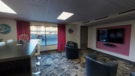 This single-floor space offers a vibrant and thoughtfully designed interior that blends comfort with bold style. The room is anchored by a patterned carpet featuring sweeping grey and black tones that add a dynamic visual energy underfoot. Seating is centered around a pair of navy blue tub chairs with studded detailing, arranged around a small accent table — ideal for intimate conversation or relaxed lounging. A large flat-screen television is mounted on the wall within a pink-framed accent panel, serving as a focal point for entertainment. Striking crimson curtains frame a wide picture window that allows natural light to fill the space while offering views of the surrounding exterior. A counter-height bar ledge with white swivel stools lines the window wall, providing an additional seating option. The ceiling features recessed grid paneling with an integrated flat panel light fixture, contributing to the room's clean, finished look. A decorative floral wall clock and a neutral-toned door round out the space's functional yet stylish character. The overall ambiance is lively and contemporary, with the color palette of navy, crimson, and pink creating a bold, welcoming atmosphere suited for social gatherings or leisure use.