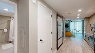 This single-floor space showcases a thoughtfully designed modern interior with a cohesive neutral palette throughout. Light wood-grain luxury vinyl flooring runs continuously across all areas, unifying the layout and lending a warm, contemporary feel.

The hallway introduces a standout architectural element: a sleek sliding barn door with a dark metal frame and frosted glass panels, offering a stylish transition between spaces while allowing light to filter through. A standard interior door with matte black hardware provides access to additional rooms.

The living area features exposed ceiling beams painted in a deep charcoal tone, adding an industrial contrast to the soft grey walls. Large windows invite natural light, and the space is open enough to accommodate comfortable seating and a tall fiddle-leaf fig plant that adds organic texture.

The kitchen is well-appointed with floor-to-ceiling wood-grain cabinetry, matte black hardware, a subway tile backsplash with under-cabinet lighting, a stainless steel range and refrigerator, and a built-in sink with a matte black faucet — all combining functionality with modern aesthetics.

The bathroom features large-format light tile flooring, a soaking tub with a curved rod and curtain, warm wood vanity cabinetry, and neutral walls, creating a clean and calming atmosphere.