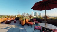 This expansive rooftop terrace is a thoughtfully designed outdoor amenity space that provides an exceptional setting for both relaxation and social gatherings. The terrace is finished with large-format concrete pavers throughout, giving the space a clean, contemporary aesthetic that complements the surrounding urban environment.

Multiple distinct seating zones are arranged across the terrace, each catering to different uses. A generous lounge area features a curved sectional sofa with vibrant, multicolored cushions, anchored by a large shade umbrella and accent tables — perfect for casual conversation or unwinding. Additional lounge chairs with bold orange cushions are positioned to take full advantage of the sweeping city skyline views visible across the roofline.

Tall, statement planters filled with ornamental grasses, seasonal flowers, and lush greenery are strategically placed throughout the terrace, adding color, texture, and a sense of privacy between seating areas. The planting design brings a garden-like quality to the rooftop setting.

An outdoor dining area offers multiple bistro-style tables with coordinating striped cushion chairs, all shaded by deep red market umbrellas. Adjacent to this area, a fully equipped outdoor kitchen stations multiple gas grills alongside a prep sink, making al fresco cooking both convenient and practical. The combination of open sky, panoramic city views, and well-appointed amenities makes this rooftop terrace a standout communal destination.