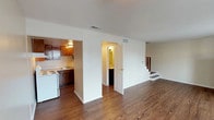 This two-floor space is designed with a practical, straightforward layout that maximizes functionality across both levels. The main floor features rich, dark hardwood-style flooring that extends throughout the open living area, creating a warm and cohesive aesthetic. A staircase with a white railing connects the two floors, positioned along one wall to preserve the openness of the main living space. The kitchen is tucked adjacent to the living area and is equipped with warm-toned wood cabinetry, a double-basin stainless steel sink, light-colored countertops, and a white electric range. A full bathroom on the main floor includes a tiled tub-and-shower combination with decorative tile accents, hardwood-style flooring, and a toilet. The upper floor transitions to neutral-toned carpeting, providing a softer feel suited to bedroom spaces. These rooms are bright and simply designed, each featuring windows with blinds that allow natural light to filter in. The overall palette throughout the space is neutral, with beige and white tones on the walls complementing the flooring choices on each level, creating a clean and understated atmosphere.