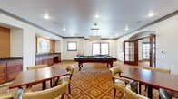 This single-floor community gathering space is designed to encourage socializing and leisure through a thoughtfully arranged series of interconnected rooms. A richly patterned carpet with a floral medallion motif flows throughout, tying the various zones together with a warm, cohesive aesthetic.

The central highlight is a generously sized billiards room, anchored by a full-size pool table beneath a pendant light fixture. Natural light enters through multiple windows, while a built-in wet bar with dark granite countertops and warm wood cabinetry adds convenient functionality. Multiple seating tables with upholstered chairs are arranged around the room for card games or casual gatherings.

Adjacent to the billiards area, a comfortable lounge features a tiled fireplace framed by a terracotta-toned surround and a framed artwork above the mantle. Cushioned armchairs and accent tables create an intimate seating arrangement, while French doors open to an outdoor balcony, inviting natural light and outdoor views.

A library and activity room provides built-in wooden bookshelves stocked with materials, alongside tables and chairs suited for reading or group activities. Framed equestrian artwork adorns the walls throughout, reinforcing a classic, refined character. Crown molding, recessed lighting, and warm-toned walls complete the inviting, well-appointed ambiance of this community space.