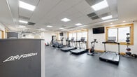 Located at 1855 Saint Francis St in Reston, VA, this fitness center offers a comprehensive workout facility within a single, open-plan space. The room is finished with durable interlocking rubber flooring in a neutral gray tone, providing a practical and comfortable surface for a wide range of exercises. A suspended drop ceiling with recessed panel lighting ensures bright, even illumination throughout the space.

Large wood-trimmed windows line the walls, allowing abundant natural light to fill the room while offering views of the surrounding area. Multiple wall-mounted flat-screen TVs are positioned throughout for entertainment during workouts.

The cardio section is well-appointed with a row of Life Fitness treadmills, elliptical cross-trainers, and a stationary spin bike, accommodating a variety of aerobic training preferences. A dedicated strength training area features a multi-station cable machine, a selectorized weight machine, an adjustable dumbbell rack with a full range of free weights, a weight bench, and various accessories including stability balls and resistance tools. A mirrored wall in the strength zone enhances the sense of space and aids in form monitoring. A beverage vending machine is also available on-site for added convenience.