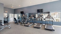 Located at 9760 Ashley River Dr. in Etna, Ohio, this fitness center offers a thoughtfully designed, single-floor workout facility. The space is finished in a modern aesthetic, featuring bold slate-blue accent walls, durable speckled rubber flooring, and recessed lighting that creates a bright, energizing atmosphere throughout.

The cardio zone is prominently anchored along one wall, outfitted with multiple treadmills, elliptical machines, and a recumbent bike. A full-length mirror spans the cardio area, visually expanding the space, while wall-mounted flat-screen televisions provide entertainment during workouts. A ceiling fan contributes to air circulation and comfort.

The free weights area includes a multi-tier dumbbell rack stocked with a range of weights, a workout bench, and additional stationary cycling equipment, offering ample options for strength training. Motivational fitness artwork adorns the walls, adding an energetic visual element to the environment.

A distinct, glass-enclosed area within the space provides a semi-private zone equipped with stability balls, resistance tools, and yoga accessories, ideal for stretching or functional training. Natural light filters in through windows, keeping the space open and inviting. The facility is well-organized, cleanly maintained, and designed to support a comprehensive range of fitness routines.