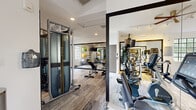 Located at the Hammocks community in Miami, FL, this fitness center offers a comprehensive workout environment within a single, open floor. The space features a modern design with crisp white walls, wood-style flooring accented with patterned carpet sections, and recessed lighting throughout that keeps the environment bright and energizing.

The cardio zone is well-appointed with multiple treadmills, elliptical trainers, and stationary bikes from Matrix, positioned near large arched and grid-pane windows that flood the area with natural light and provide views of the surrounding greenery. A wall-mounted flat-screen television accompanies this section for entertainment during workouts.

The strength training area includes a Matrix functional trainer cable machine, a multi-tier dumbbell rack with a full range of weights, kettlebells, and an adjustable workout bench. Large wall-mounted mirrors expand the sense of space and allow users to monitor their form. A dedicated stretching and functional fitness zone features stability balls, exercise mats, and additional benches, supported by its own mirrored wall and natural light from multiple windows.

Practical amenities include a built-in water fountain, a freestanding water station, and a glass double-door entry that connects directly to an outdoor area. The overall ambiance is clean, airy, and purpose-built to support a wide range of fitness activities.