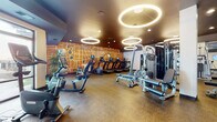 This modern fitness center is thoughtfully designed to support a full range of workout routines in a stylish and functional environment. The space is anchored by a bold, oversized map-inspired mural rendered in warm amber and white tones, serving as a striking focal point across the main wall and lending the room a distinctive urban character.

Overhead, a series of circular halo pendant lights illuminate the space with a warm, ambient glow, complemented by recessed ceiling lighting throughout. The dark-toned ceiling adds depth and contrast, while durable cork-style flooring provides a practical and resilient surface underfoot.

The cardio zone is well-stocked with treadmills, elliptical trainers, and stationary bikes, each equipped with individual digital displays. Rowing machines and additional functional training equipment round out the aerobic offerings. On the strength side, the facility includes a multi-station cable and weight machine, a full rack of dumbbells, medicine balls, stability balls, and weight benches — all positioned alongside floor-to-ceiling mirrors that help maximize the sense of space.

A built-in water fountain station is conveniently located near the free weights area. Large windows along one wall welcome natural light and offer views to the exterior, enhancing the overall atmosphere. Access to the fitness center is secured via a key-card entry system, and a glass-partitioned divider adds an open, connected feel between zones.