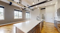 Located in the heart of downtown Hornell, NY, this two-story loft-style residence masterfully blends industrial character with modern finishes. The expansive main floor is anchored by rich, reclaimed hardwood flooring that extends throughout the open-concept living and kitchen area. Soaring ceilings are accented by bold, dark-stained exposed beams and industrial ductwork, lending an authentic loft aesthetic. Large arched windows with painted brick surrounds flood the space with natural light while offering views of the surrounding area.

The chef's kitchen is a standout feature, boasting dark shaker-style cabinetry, a white subway tile backsplash, stainless steel appliances, and a generous island with a built-in sink and pendant lighting overhead. An in-unit stacked washer and dryer add everyday convenience.

A sleek staircase with modern cable railings leads to the upper level, where dramatic angled structural beams define the space. The upper floor houses the primary bedroom suite, accompanied by a full bathroom featuring a gray vanity with white countertop and exposed beam details. The main-floor bathroom offers a tiled walk-in glass shower. Throughout, the design balances raw architectural elements with clean, contemporary finishes.