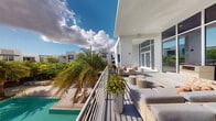 This space showcases a well-appointed outdoor terrace defined by clean, contemporary architecture with a crisp white exterior finish. The terrace runs alongside the building, offering an elevated vantage point overlooking a sparkling pool area below, framed by lush tropical palm trees and verdant landscaping that reinforce a warm, resort-inspired atmosphere.

The outdoor seating area is furnished with wicker lounge chairs and matching ottomans arranged to encourage relaxed conversation or leisure, complemented by tall cylindrical planters filled with tropical greenery that add a decorative, organic touch. A sleek metal railing lines the perimeter, providing unobstructed views of the surrounding landscape and the bright blue sky dotted with clouds above.

Adjacent to the lounge area, large glass double doors with floor-to-ceiling framing lead into an interior space, allowing for an easy indoor-outdoor flow. Recessed lighting is integrated into the covered ceiling section of the terrace, ensuring usability during evening hours. The overall ambiance is bright, airy, and modern, with the white architectural palette, natural light, and tropical surroundings combining to create a polished yet inviting outdoor environment suited for relaxation and social gatherings.