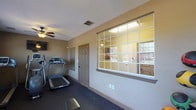 This fitness center offers a comprehensive workout environment designed to support a range of exercise routines. The space is organized into distinct functional zones, making it easy to transition between cardio and strength training. The cardio area is outfitted with multiple treadmills and an elliptical machine, each featuring digital consoles, and a wall-mounted flat-screen television provides entertainment during workouts. The strength training area includes a full dumbbell rack with a wide range of weights, a medicine ball collection, a hyperextension bench, and a weight bench, catering to users of all fitness levels. A large mirror spans one wall of the free weights area, enhancing the sense of space and allowing for proper form checks. Rubber flooring throughout provides cushioning and durability underfoot. Natural light filters in through multiple windows fitted with blinds, while ceiling fans help maintain a comfortable atmosphere. The warm, neutral-toned walls and practical layout give the space a welcoming and functional ambiance. An interior window connects the fitness center to an adjacent room, adding openness to the overall design.