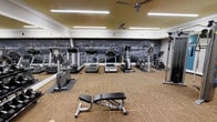 This fitness center is a single-floor facility designed to support a well-rounded workout experience. The space is outfitted with a diverse selection of cardio equipment, including treadmills, elliptical trainers, and stationary bikes, all arranged across a durable, striped low-pile carpet flooring. A weight bench positioned in the center of the room provides a dedicated area for strength training exercises. The room is brightly lit with overhead fluorescent lighting, while two ceiling fans promote air circulation and comfort during workouts. A full-length mirror along one wall enhances the sense of space and allows users to monitor their form. The most striking design element is the large-scale black-and-white mountain landscape mural that spans the length of the back wall, set against a bold dark background with a warm golden-yellow upper wall accent, creating an energizing and visually dynamic atmosphere. White crown molding and trim add a clean, finished look to the interior. The overall ambiance is functional yet thoughtfully designed, offering an inviting environment that motivates users while providing all the essential equipment for a comprehensive fitness routine.