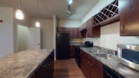 This single-floor residence offers a thoughtfully designed open-concept layout that seamlessly connects the living and kitchen areas. The kitchen is a standout feature, equipped with rich dark wood cabinetry, granite-style countertops, black appliances including a gas range and refrigerator, a built-in wine rack, and a central island with pendant lighting overhead. Track lighting enhances the workspace, while hardwood-style flooring runs throughout the main living areas, adding warmth and continuity.

The open living area is generously sized, offering a clean, versatile space with hardwood-style floors that extend from the kitchen. Natural light filters in through windows near the front entry, which features a ceiling fan and a welcoming foyer. Bedrooms are carpeted for comfort and receive ample natural light through large windows fitted with blinds.

The bathrooms are consistently finished with dark wood vanities, granite-style countertops, single basin sinks, large mirrors, and full soaking tub and shower combinations with tiled or paneled surrounds. Hardwood-style flooring carries into the bathrooms as well, maintaining a cohesive aesthetic throughout. Overall, the space presents a clean, modern interior with a consistent design language across all rooms.
