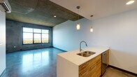 Located at 122 Delaware St in Kansas City's vibrant River Market neighborhood, this contemporary single-floor unit showcases an industrial-modern aesthetic throughout. Polished concrete floors run seamlessly across the entire space, reflecting natural light and complementing the exposed concrete ceiling that adds raw architectural character.

The open-concept layout places the kitchen at the heart of the unit, equipped with stainless steel appliances including a full-size refrigerator, electric range, and built-in microwave. Warm wood-toned cabinetry is paired with clean white countertops on both the perimeter and the central island, which features a double stainless steel sink and stylish pendant lighting overhead. White upper cabinets provide ample storage.

The expansive living area benefits from a large picture window that frames sweeping views of the surrounding greenery and cityscape, flooding the room with natural light. A wall-mounted mini-split unit ensures year-round climate control.

The bathroom features a sleek vanity with a white countertop, wood cabinetry, and a large mirror, with a stacked washer and dryer conveniently located nearby for in-unit laundry. The overall palette of soft blue-gray walls, concrete, and natural wood tones creates a cohesive, contemporary ambiance throughout this well-appointed urban residence.