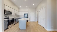 This single-floor residence offers a thoughtfully designed layout with contemporary finishes throughout. The kitchen serves as a central highlight, featuring white shaker-style upper cabinetry paired with gray lower cabinets, light quartz countertops, and a full suite of stainless steel appliances including a range, microwave, and dishwasher, complemented by a black refrigerator. A gray center island with a white countertop provides additional prep space and separates the kitchen from the adjoining living area, which benefits from an open, airy feel enhanced by recessed lighting and elevated ceilings. Light wood-look flooring flows consistently through the main living spaces, adding warmth to the neutral color palette of white and soft gray tones. The bathroom features a substantial gray vanity with ample drawer storage, a white countertop, and a well-lit mirror. The bedroom is carpeted for comfort and includes two windows with blinds that bring in natural light. A dedicated laundry area houses a full-size top-load washer and dryer, providing added convenience. The overall design is clean, modern, and cohesive.