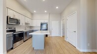 This single-floor residence offers a thoughtfully designed layout with contemporary finishes throughout. The kitchen serves as a central highlight, featuring white shaker-style upper cabinetry paired with gray lower cabinets, light quartz countertops, and a full suite of stainless steel appliances including a range, microwave, and dishwasher, complemented by a black refrigerator. A gray center island with a white countertop provides additional prep space and separates the kitchen from the adjoining living area, which benefits from an open, airy feel enhanced by recessed lighting and elevated ceilings. Light wood-look flooring flows consistently through the main living spaces, adding warmth to the neutral color palette of white and soft gray tones. The bathroom features a substantial gray vanity with ample drawer storage, a white countertop, and a well-lit mirror. The bedroom is carpeted for comfort and includes two windows with blinds that bring in natural light. A dedicated laundry area houses a full-size top-load washer and dryer, providing added convenience. The overall design is clean, modern, and cohesive.