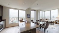 This contemporary high-rise residence offers a refined urban living experience defined by clean lines, modern finishes, and expansive city views. The open-concept living, dining, and kitchen area is anchored by dramatic floor-to-ceiling windows on multiple walls, flooding the space with natural light and framing panoramic skyline vistas. The kitchen is outfitted with dark, sophisticated cabinetry, a large central island with an integrated sink, and premium stainless steel Bosch appliances including a wall oven, induction cooktop, and over-the-range microwave. Light hardwood flooring flows throughout the main living areas, complementing the neutral, contemporary palette. The spacious living room features a corner arrangement of floor-to-ceiling glass, maximizing the sweeping city outlook. The primary bedroom is a serene retreat with its own en-suite bath, which boasts an elongated white vanity with LED-lit mirror, large-format tile flooring, and a separate shower. A secondary bedroom comfortably accommodates twin beds with its own window framing elevated city views. A second full bathroom mirrors the same clean, modern aesthetic. Throughout, the residence maintains a cohesive design language of bright whites, warm wood tones, and polished surfaces.