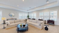 This thoughtfully designed single-floor space combines contemporary style with functional comfort across its open, airy layout. Light wood-look flooring runs throughout, unifying the distinct areas and enhancing the clean, modern aesthetic.

The spacious lounge area serves as the heart of the space, anchored by a generously sized sectional sofa and a round dark wood coffee table. Elegant tray ceilings with recessed lighting add architectural depth, while wainscoting along the lower walls lends a refined touch. Large windows fitted with white horizontal blinds line multiple walls, flooding the room with natural light. A separate seating nook along the window wall features cushioned barrel chairs and small side tables, creating an intimate setting for conversation or relaxation.

The kitchen is both stylish and well-appointed, featuring light wood cabinetry with matte black hardware, white quartz countertops, and stainless steel appliances including a full-size refrigerator and built-in oven. A substantial island with a dark wood base and white countertop provides casual seating for several guests, with globe pendant lights overhead. A double stainless steel sink sits beneath a bright window, completing a practical yet attractive workspace.

The overall ambiance is calm and welcoming, with a neutral color palette, modern finishes, and an abundance of natural light creating an inviting environment throughout.