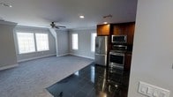 This single-floor space showcases an open-concept layout that seamlessly connects the kitchen and living area, creating a spacious and functional environment. The kitchen is anchored by rich, warm-toned wood cabinetry that extends to the ceiling, providing ample storage. A suite of stainless steel appliances — including a French door refrigerator, a double wall oven, and a microwave — lends a modern, professional feel to the cooking space. The kitchen floor features glossy black tiles that add a bold, polished contrast to the surroundings.

The adjacent living area transitions to neutral-toned carpeting, offering a softer, comfortable surface underfoot. Light gray walls throughout the space provide a clean, versatile backdrop, while white crown molding and baseboards add a refined architectural detail. Multiple windows along the walls allow natural light to filter in generously, brightening the interior. A ceiling fan with a light fixture is centrally positioned in the living area, providing both air circulation and additional lighting. Recessed lighting in the kitchen area completes the well-considered lighting scheme, making this an inviting and practical space overall.