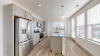 Located at 210 W Fifth Ave in Columbus, Ohio, this single-floor residence offers a clean, contemporary design with a bright and airy atmosphere throughout. The open-concept kitchen is a standout feature, showcasing soft gray shaker-style cabinetry extending to the ceiling for ample storage, granite countertops, and a central island with a built-in sink and pendant lighting above. Stainless steel appliances, including a French door refrigerator, electric range, and over-the-range microwave, complete the well-appointed cooking space. Large windows dressed with blinds invite an abundance of natural light into both the kitchen and the adjacent living area, which features light hardwood-style flooring that runs seamlessly throughout the main living spaces. The spacious living room offers a generous, open layout ideal for a variety of configurations. The bedroom is carpeted for comfort and includes access to a walk-in closet outfitted with wire shelving systems for organized storage. The bathroom features a walk-in shower with a glass sliding door and built-in shelving. The consistent use of neutral tones, recessed lighting, and clean architectural lines gives the entire space a fresh, move-in-ready appeal.