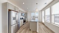 Located at 210 W Fifth Ave in Columbus, Ohio, this single-floor residence offers a clean, contemporary design with a bright and airy atmosphere throughout. The open-concept kitchen is a standout feature, showcasing soft gray shaker-style cabinetry extending to the ceiling for ample storage, granite countertops, and a central island with a built-in sink and pendant lighting above. Stainless steel appliances, including a French door refrigerator, electric range, and over-the-range microwave, complete the well-appointed cooking space. Large windows dressed with blinds invite an abundance of natural light into both the kitchen and the adjacent living area, which features light hardwood-style flooring that runs seamlessly throughout the main living spaces. The spacious living room offers a generous, open layout ideal for a variety of configurations. The bedroom is carpeted for comfort and includes access to a walk-in closet outfitted with wire shelving systems for organized storage. The bathroom features a walk-in shower with a glass sliding door and built-in shelving. The consistent use of neutral tones, recessed lighting, and clean architectural lines gives the entire space a fresh, move-in-ready appeal.
