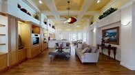 The Quantum Lakes Clubhouse in Boynton Beach, Florida, is a thoughtfully designed single-story community facility that blends classic architectural details with comfortable, functional interiors. The main gathering area is immediately distinguished by soaring coffered ceilings with warm yellow accents and crisp white crown molding, creating a grand yet welcoming atmosphere. A decorative ceiling fan serves as a focal point overhead, complementing the recessed lighting throughout.

Rich hardwood flooring runs continuously across the space, anchoring the various seating and activity zones. The main lounge features generously sized neutral sofas arranged around an ornate glass-top coffee table, with built-in wood cabinetry and open shelving along one wall housing a television and decorative accents. Tall decorative columns and wall sconces add architectural character throughout.

A curved peninsula counter with granite surfaces and warm wood upper cabinetry defines the kitchenette area, suitable for community gatherings and events. Adjacent to it, a sunlit dining area offers multiple glass-top tables with traditional wood chairs, bathed in natural light from large arched and grid-pane windows overlooking the outdoor courtyard. Lush tropical plants are positioned throughout, reinforcing the Florida setting and adding vibrancy to the overall ambiance.