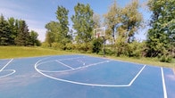 This outdoor basketball court offers a well-maintained recreational space designed for active play and community engagement. The court features a striking blue sport surface with clearly marked white boundary lines, a center circle, and a key area, all indicating a regulation-style layout suited for both casual games and organized play.

A sturdy steel pole supports a full-sized backboard with a standard rim and net, providing a professional-grade setup for players of all skill levels. The court is set at ground level, seamlessly integrated into the surrounding landscape.

The natural environment surrounding the court is one of its most appealing characteristics. Lush, mature trees frame the perimeter, providing partial shade and a scenic green backdrop. An open grassy area adjoins the court, offering additional space for spectators or warm-up activities.

Under open skies, the court benefits from abundant natural light, creating an inviting atmosphere on clear days. The combination of the vibrant playing surface, quality equipment, and verdant surroundings makes this an attractive and functional outdoor athletic space, well-suited for recreation and physical activity in a natural setting.