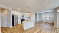 Located in Forest Village, Holly Springs, NC, this single-floor residence offers a thoughtfully designed open-concept layout filled with natural light. The kitchen is a standout feature, boasting white shaker-style cabinetry, granite countertops, a herringbone tile backsplash, and a full suite of stainless steel appliances including a refrigerator, range, and over-the-range microwave. A central island with a undermount sink and pendant lighting above serves as both a functional workspace and a gathering point that flows seamlessly into the living area. Warm wood-look luxury vinyl plank flooring extends throughout the main living spaces, complementing the neutral color palette and clean lines of the interior. The living area is bright and open, with windows fitted with blinds and access to an exterior door. Bedrooms are carpeted for comfort, with ceiling fans, natural light, and closets featuring built-in rods for storage. The bathrooms are well-appointed with granite-topped vanities, undermount sinks, large mirrors, and wood-look flooring. A dedicated closet space and accessible laundry area add to the unit's practical appeal. The overall ambiance is clean, contemporary, and move-in ready.