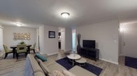 This single-floor residence offers a well-organized layout across multiple interconnected living spaces, finished with a consistent neutral color palette and wood-look flooring throughout the main areas. The living room is generously sized, featuring a large sliding glass door dressed with vertical blinds that floods the space with natural light and provides outdoor views. A wall-mounted air conditioning unit and baseboard heating are also present, ensuring year-round comfort.

Adjacent to the living area, a dedicated dining space accommodates a round wooden table and connects directly to the kitchen. The kitchen is outfitted with dark espresso cabinetry, granite-style countertops, and black appliances including a refrigerator, electric range, and overhead ventilation hood, all set on wood-look flooring.

The unit includes two bedrooms, both carpeted for warmth and comfort. Each room features vertical-blind-covered windows that allow natural light to filter in. One bedroom includes a sliding closet, providing practical storage. The bathroom is cleanly finished with a modern floating vanity, a full tub-shower combination, and chrome fixtures throughout, maintaining the home's cohesive, contemporary aesthetic.