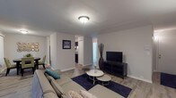 This single-floor residence offers a well-organized layout across multiple interconnected living spaces, finished with a consistent neutral color palette and wood-look flooring throughout the main areas. The living room is generously sized, featuring a large sliding glass door dressed with vertical blinds that floods the space with natural light and provides outdoor views. A wall-mounted air conditioning unit and baseboard heating are also present, ensuring year-round comfort.

Adjacent to the living area, a dedicated dining space accommodates a round wooden table and connects directly to the kitchen. The kitchen is outfitted with dark espresso cabinetry, granite-style countertops, and black appliances including a refrigerator, electric range, and overhead ventilation hood, all set on wood-look flooring.

The unit includes two bedrooms, both carpeted for warmth and comfort. Each room features vertical-blind-covered windows that allow natural light to filter in. One bedroom includes a sliding closet, providing practical storage. The bathroom is cleanly finished with a modern floating vanity, a full tub-shower combination, and chrome fixtures throughout, maintaining the home's cohesive, contemporary aesthetic.