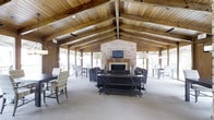 This two-story space makes an immediate impression with its soaring vaulted ceiling, defined by rich exposed wooden beams and tongue-and-groove wood planking that lend the interior a warm, lodge-inspired character. The main gathering area is anchored by a classic brick fireplace fitted with a traditional mantel and a mounted flat-screen television, serving as a natural focal point for the room. Generous seating arrangements, including plush sofas and accent chairs, are thoughtfully arranged around the fireplace to encourage conversation and relaxation. A substantial X-frame console table adds an industrial-rustic touch to the central space. Floor-length windows line the perimeter of the room, flooding the interior with abundant natural light and offering views of the surrounding outdoor areas, while also providing access to what appears to be an exterior deck or patio. Dining and casual seating areas are positioned near the windows, making them ideal for both social gatherings and quiet retreat. The neutral carpeting and muted tones of the furnishings complement the natural wood and brick elements throughout, creating a cohesive and inviting ambiance suited to a wide range of uses across both floors.