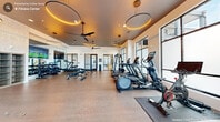 Located in Cary, NC, this contemporary fitness center offers a well-equipped, single-floor workout facility designed with both function and aesthetics in mind. The space features a clean, open layout with durable wood-look flooring throughout, providing an inviting environment for a full range of exercise activities.

Cardio equipment is prominently featured along the windowed wall, including treadmills, ellipticals, a stair climber, and Peloton spin bikes, all positioned to take advantage of abundant natural light streaming through large, floor-to-ceiling windows. A dedicated free weights area includes a full rack of dumbbells alongside adjustable benches, with a large wall-mounted mirror and flat-screen TV enhancing the workout experience. A cable and strength machine station rounds out the equipment offerings, supporting a comprehensive full-body training routine.

The ceiling is a notable architectural highlight, showcasing dramatic circular LED ring pendant lights that cast a warm, amber glow throughout the space. Ceiling fans complement the lighting fixtures, contributing to comfort during intense workouts. Textured accent walls and built-in shelving add refined design touches, while glass-paneled entry doors provide a sleek, modern entrance. The overall ambiance is polished and motivating, combining upscale design elements with practical, high-quality fitness amenities.