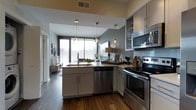 This single-floor space showcases a thoughtfully designed, open-concept kitchen and living area. The kitchen is outfitted with stainless steel appliances, including a smooth-top electric range, built-in microwave, and dishwasher. White shaker-style cabinetry with sleek hardware lines the walls, complemented by upper glass-front cabinets that add visual depth. Light countertops provide ample workspace, while a peninsula with an undermount sink serves as both a functional prep area and a natural divider between the kitchen and living space. Three glass pendant lights hang above, adding a refined touch to the layout. Dark hardwood flooring runs throughout, grounding the space with warmth and contrast against the light cabinetry and gray-toned walls. A subtle tile backsplash ties the kitchen's design elements together. The space opens toward a bright living area, where floor-to-ceiling windows allow generous natural light to filter through, creating an airy and inviting atmosphere. The overall aesthetic is contemporary and cohesive, blending functional design with modern finishes throughout.