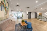 This thoughtfully designed single-floor amenity space presents a cohesive, contemporary aesthetic across all of its key areas. The welcoming entrance lobby sets the tone with large-format light stone-effect tile flooring, glass-railed stairwell access, recessed lighting, and vibrant artwork adorning the crisp white walls. A glass partition wall adds a sense of openness while defining functional zones within the corridor.

The lounge area is a standout feature, furnished with plush, modern sofas in muted tones and complemented by a richly patterned area rug. A curated gallery wall of abstract circular artworks adds a sophisticated visual focal point, while layered lighting creates a warm and inviting atmosphere ideal for gathering or relaxing.

Adjacent seating nooks continue the refined aesthetic, with additional bold abstract canvases, floor lamps, and comfortable seating that invite residents or guests to linger.

The communal kitchen and dining area is both functional and elegant, featuring white cabinetry, stainless steel appliances including a full-size refrigerator, and light tile flooring. A large wood dining table surrounded by sleek cream chairs seats a generous number of guests. Glass doors open directly onto an outdoor deck, flooding the space with natural light and providing seamless indoor-outdoor connectivity, with views of a furnished terrace beyond.