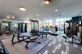 Located at 368 Sublime Summer Lane in Clover, SC, this contemporary fitness center offers a spacious, single-floor layout divided into two distinct workout zones designed to support a full range of exercise modalities. The main strength and cardio area features patterned carpet tile flooring, large floor-to-ceiling windows that flood the space with natural light and offer views of the surrounding greenery. This zone is equipped with a multi-station cable machine, adjustable weight benches, a full dumbbell rack, and multiple treadmills, providing comprehensive options for both resistance training and cardiovascular workouts. Full-length mirrors line portions of the walls, enhancing the sense of openness while allowing users to monitor their form. A second dedicated studio room features smooth hardwood-style flooring and a textured tile accent wall, anchored by a large flat-screen TV for guided workout programming. This area houses cycling bikes, a rowing machine, TRX suspension training benches, and interactive fitness equipment, making it ideal for group or tech-driven training sessions. A glass partition wall connects the two zones while maintaining visual continuity throughout the facility. Recessed and flush-mount ceiling lighting, combined with abundant window light, creates a bright and energizing atmosphere throughout.